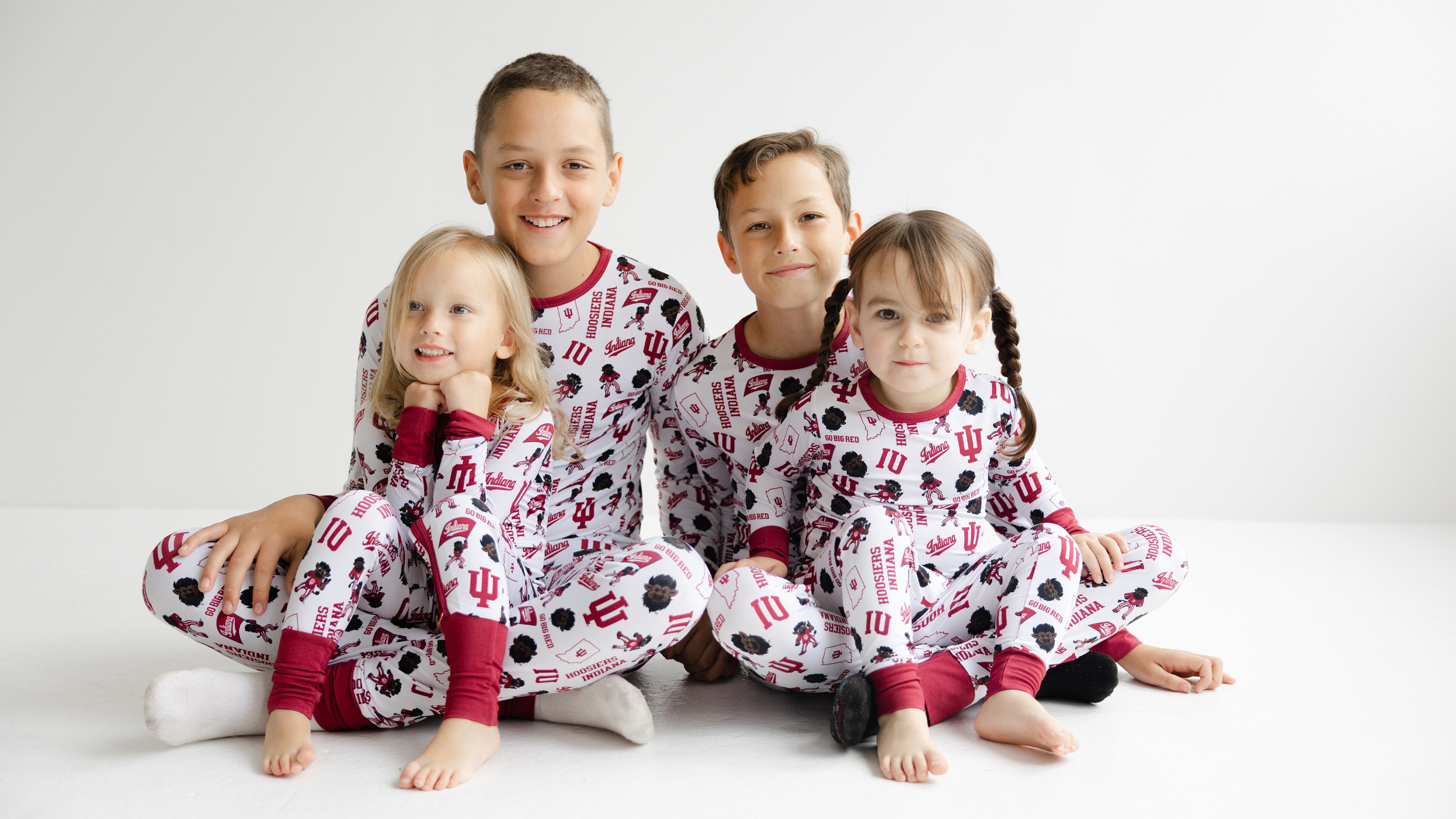 Four children sitting together wearing matching indiana hoosiers pajamas printed with red and black football motifs and red cuffs, against a clean white studio background.