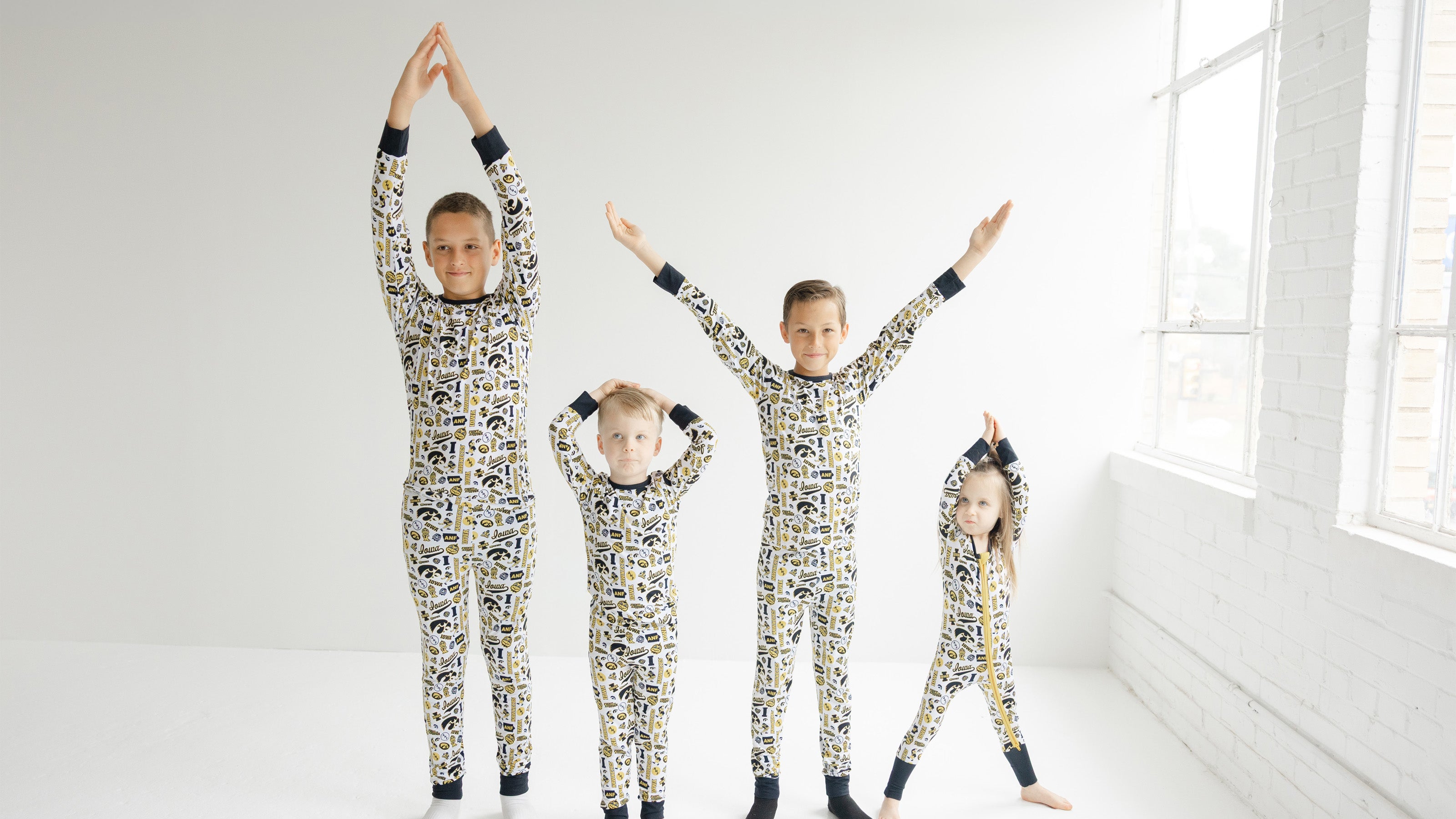 Four children wearing matching iowa hawkeyes bamboo long-sleeve pajamas with a black, white, and yellow graphic pattern, standing in a bright white room with their arms raised.