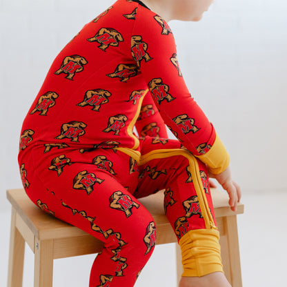 A child wearing a bright red long-sleeve pajama set with an allover Maryland terrapins Testudo mark  pattern, sitting on a light wood stool against a white background.