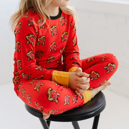 A child wearing a bright red long-sleeve pajama top and matching pants with an allover Maryland terrapins Testudo print, sitting cross-legged on a black stool against a light neutral background.