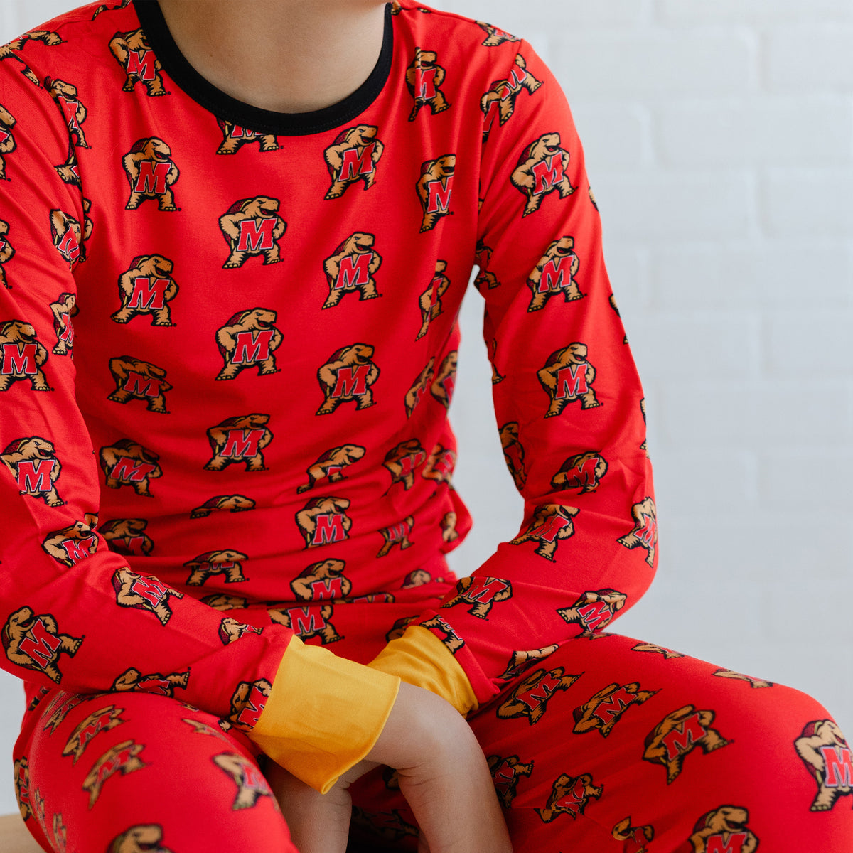 A youth wearing a red long-sleeve pajama top and matching pants with an allover Maryland terrapins Testudo print, featuring a black crew neck and a yellow inner cuff.