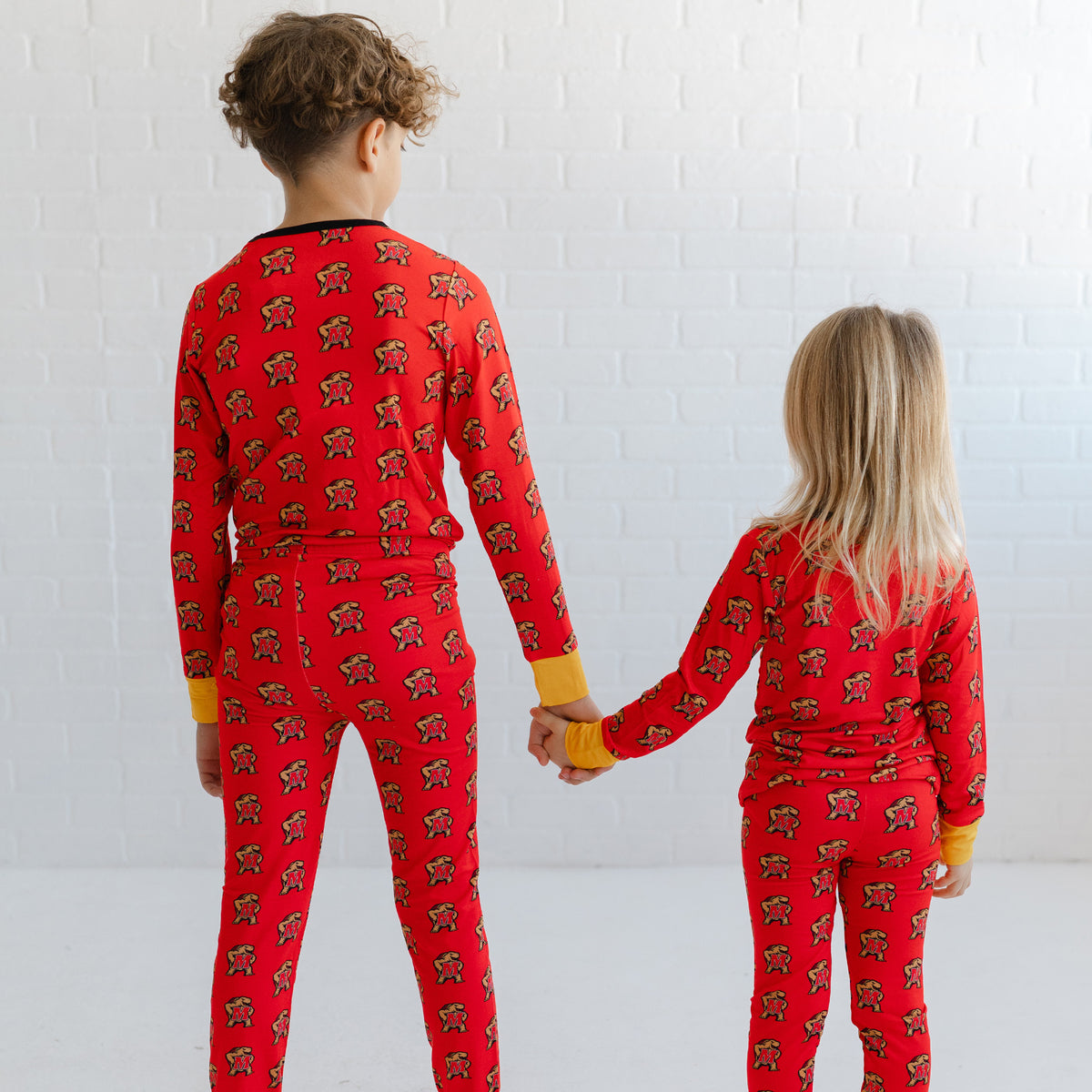 Two children standing with their backs to the camera, holding hands and wearing matching red long-sleeve pajamas with a repeating Maryland terrapins Testudo print.
