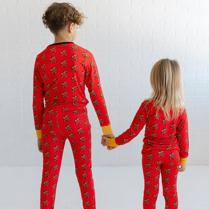 Two children standing with their backs to the camera, holding hands and wearing matching red long-sleeve pajamas with a repeating Maryland terrapins Testudo print.