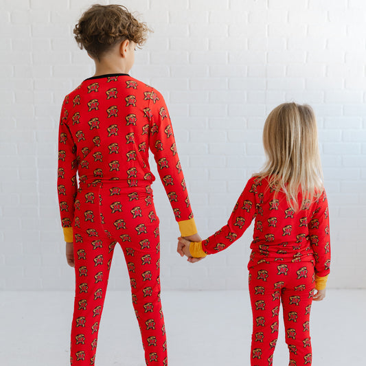Two children standing with their backs to the camera, holding hands and wearing matching red long-sleeve pajamas with a repeating Maryland terrapins Testudo print.