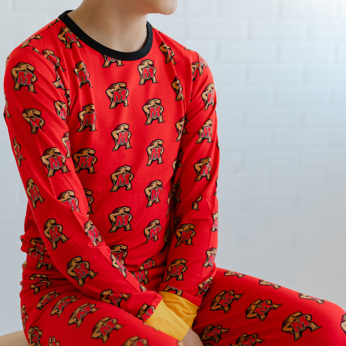 A youth wearing a bright red long-sleeve pajama top and matching pants with an allover Maryland terrapins Testudo print, seated against a light gray brick wall.