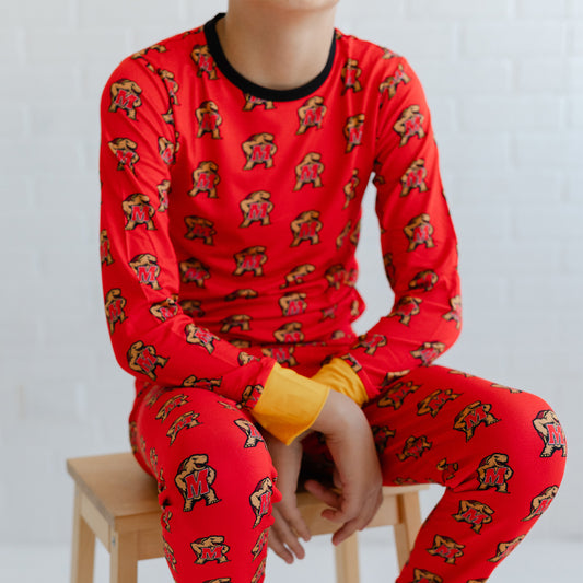 A child wearing a bright red long-sleeve pajama top and matching pants with an allover Maryland terrapins Testudo print, seated on a wooden stool against a white brick wall.