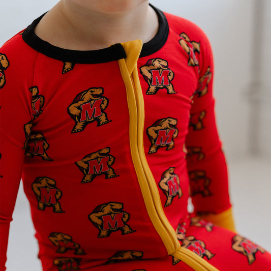 A close-up of a child wearing red long-sleeve pajamas with a black collar, yellow zipper, and an allover print of the maryland terrapins.