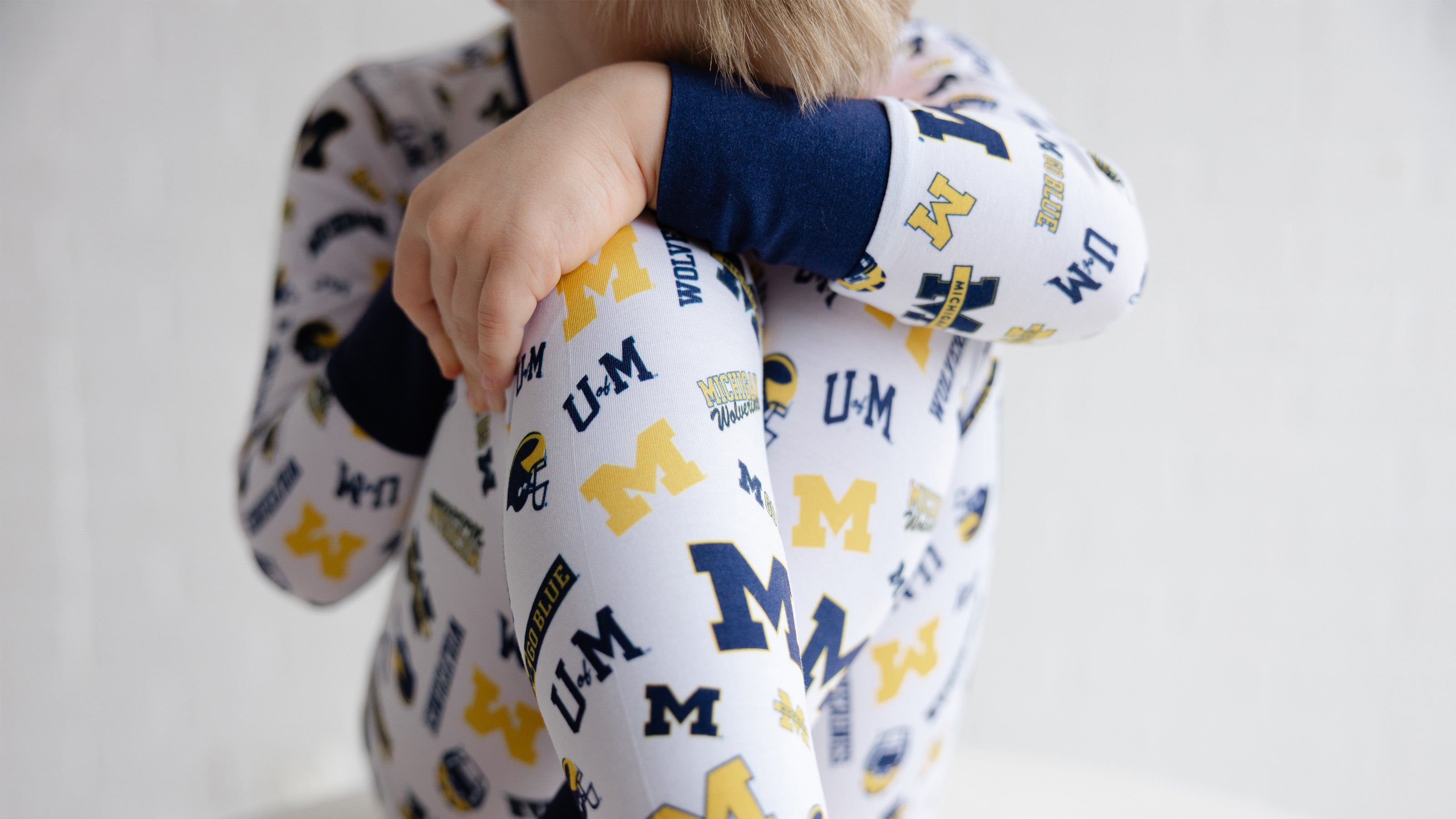 A child wearing a white long-sleeve pajama top with navy and yellow Michigan Wolverines collegiate lettering, shown from the back with arms crossed.