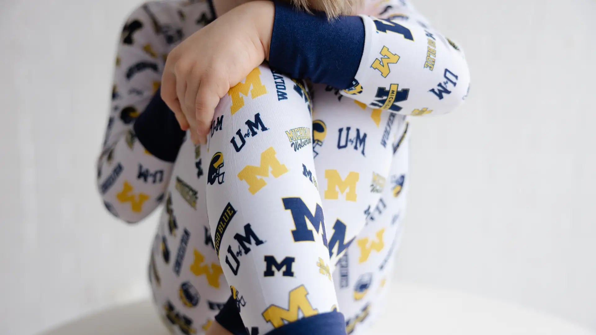 A child wearing a white long-sleeve pajama top and matching leggings with a navy and yellow Michigan Wolverines logo print, hugging their knees against a plain white background.