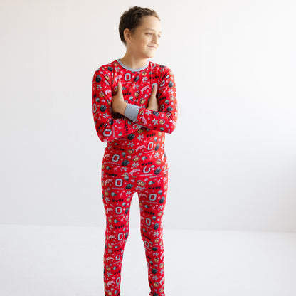 A child wearing a red long-sleeve pajama set with an allover officially licensed ohio state buckeyes bamboo pajama print, standing against a plain white studio background.