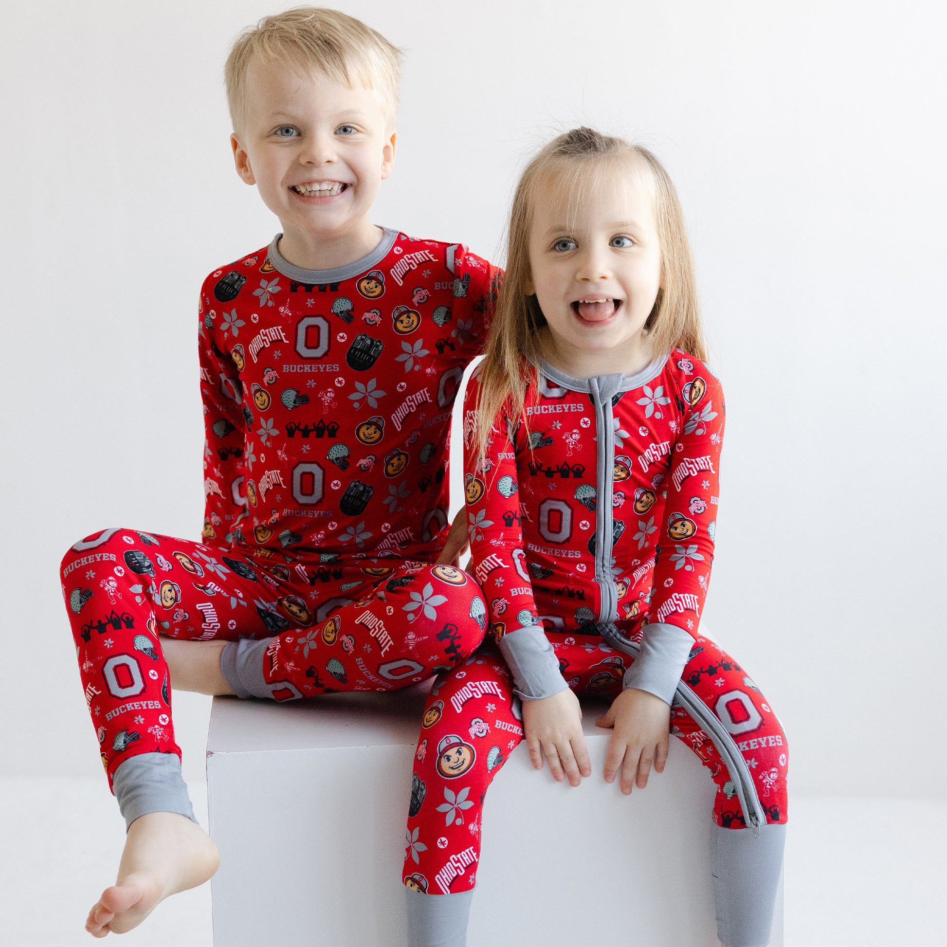Two children sitting on a white block wearing matching officially licensed ohio state buckeyes bamboo pajamas with gray cuffs and collars, photographed against a plain light background.