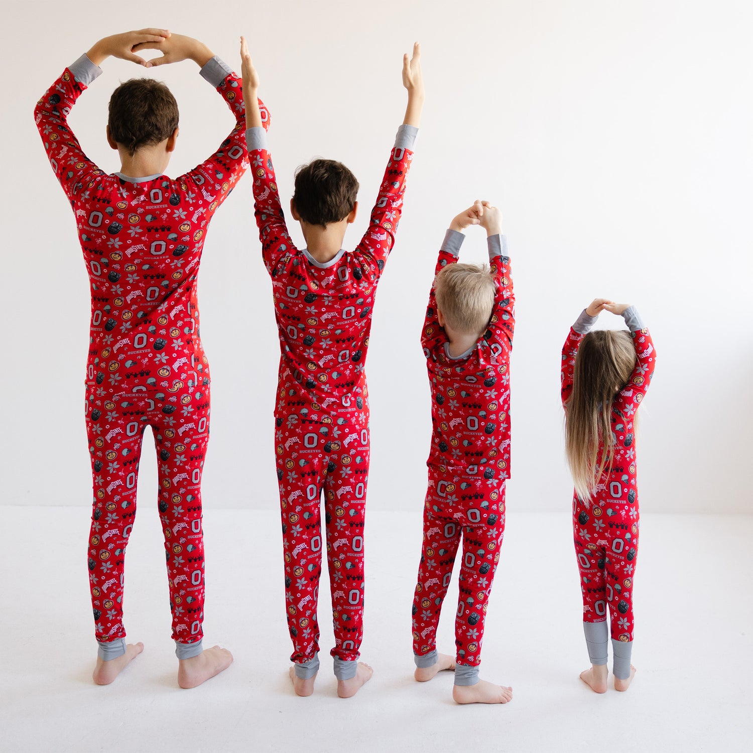 Four children standing with arms raised, wearing officially licensed ohio state buckeyes pajamas with gray cuffs and ankles, photographed against a plain white studio background.