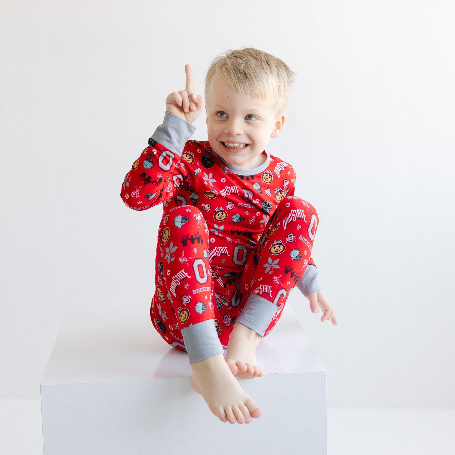 A smiling young child wearing officially licensed ohio state buckeyes with gray cuffs, sitting on a white block against a plain white background.