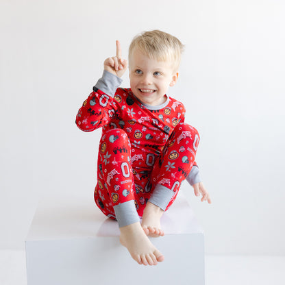 A smiling young child wearing officially licensed ohio state buckeyes with gray cuffs, sitting on a white block against a plain white background.