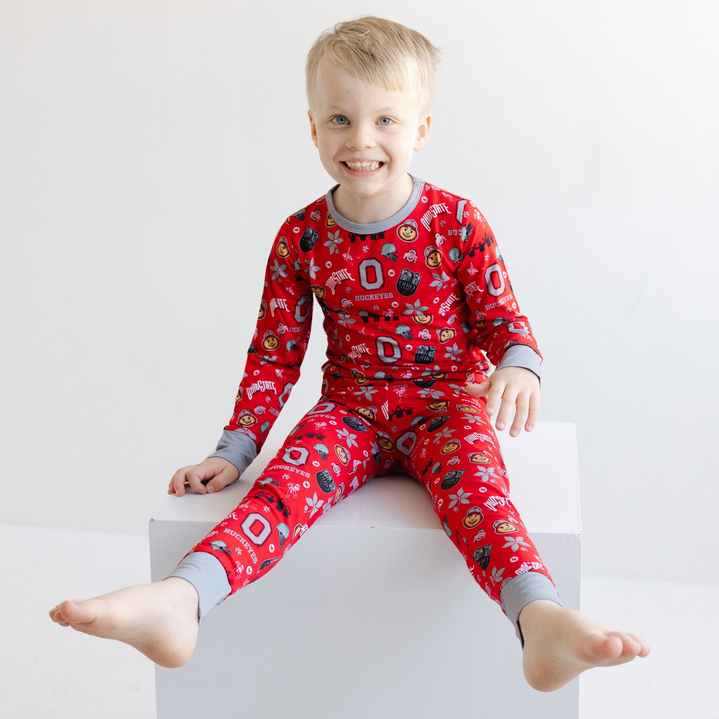 A young child wearing a red long-sleeve officially licensed ohio state buckeyes bamboo pajamas, sitting on a white block against a light gray background.