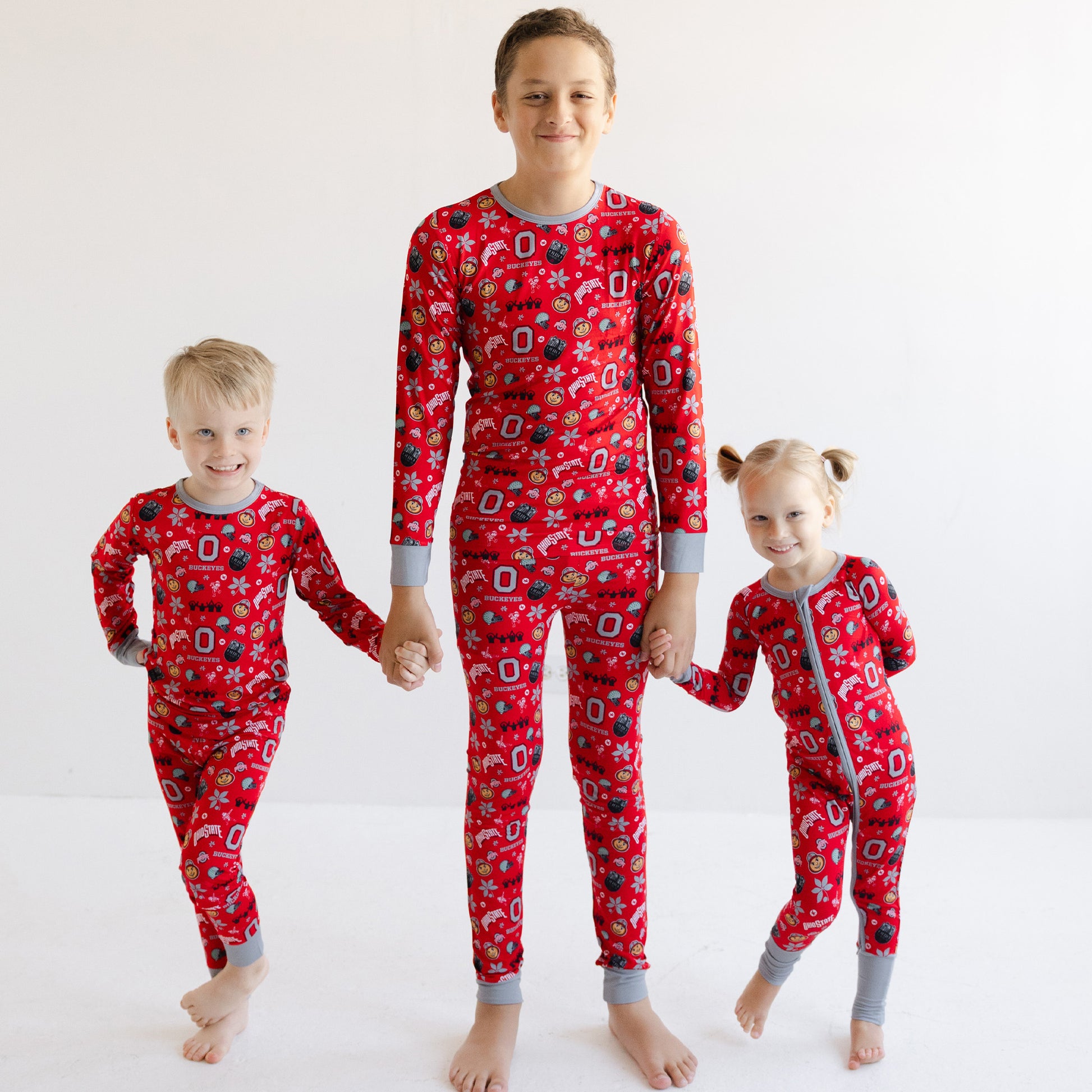 Three children standing barefoot and holding hands, wearing matching officially licensed ohio state buckeyes , set against a plain white studio background.