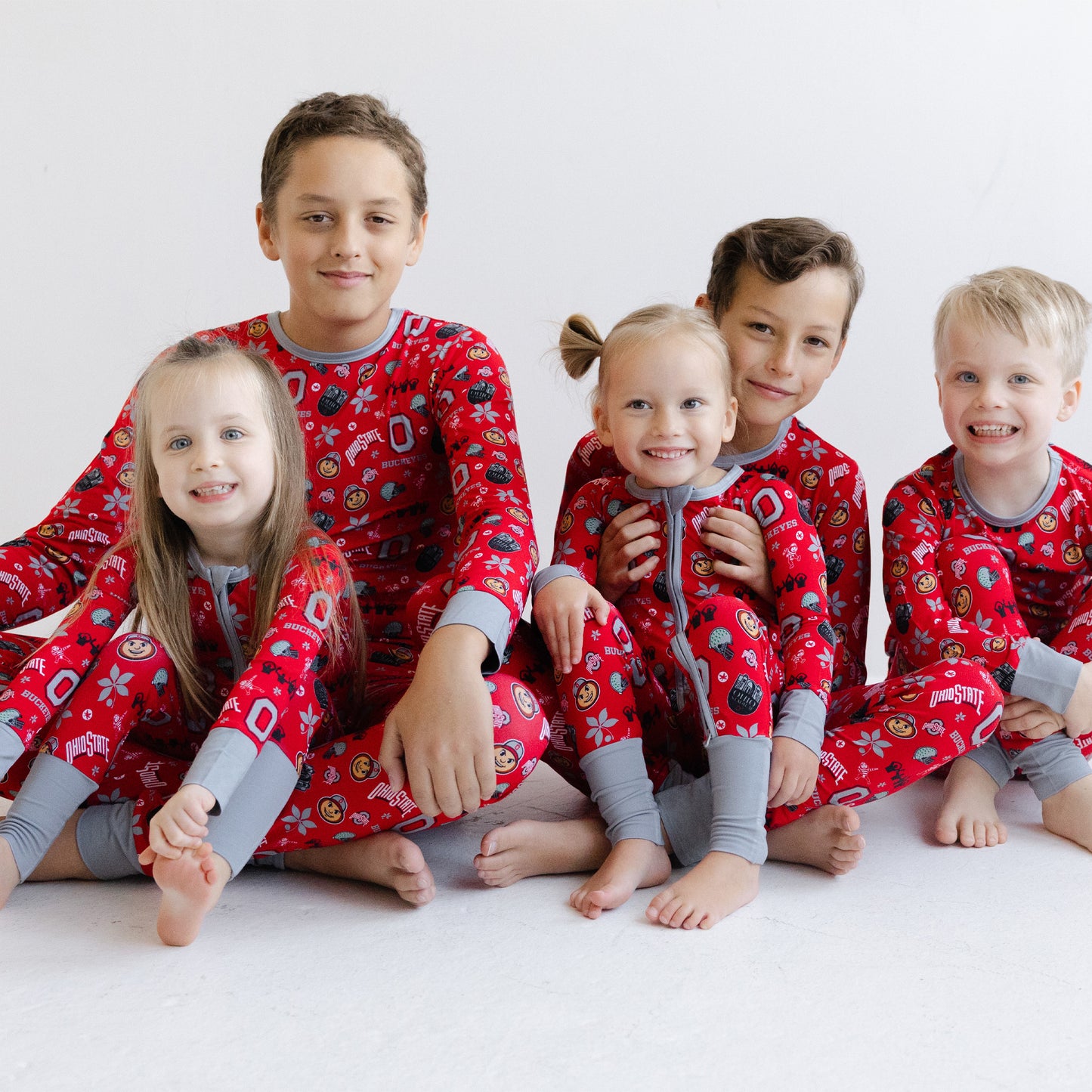 Five children sitting together wearing matching scarlet officially licensed ohio state buckeyes pajamas with gray cuffs and legs, photographed against a plain light background.