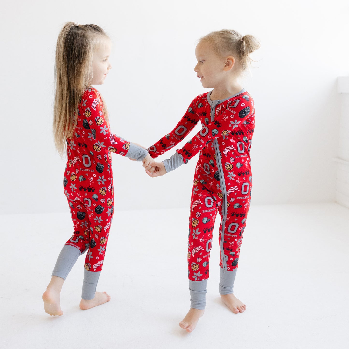 Two young girls standing and holding hands, wearing matching ohio state buckeyes pajamas with gray cuffs and collar, in a bright white room.