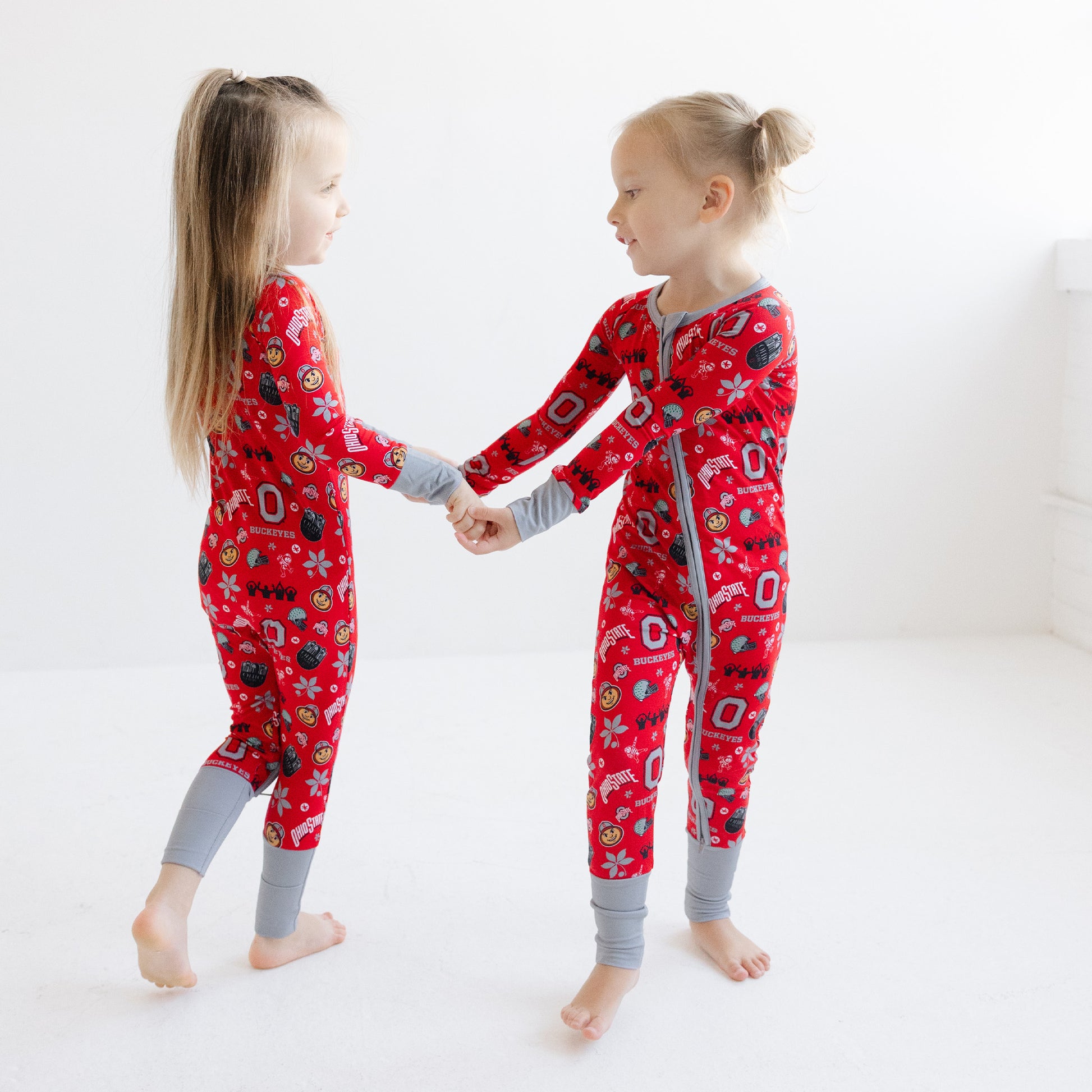 Two young girls standing and holding hands, wearing matching ohio state buckeyes pajamas with gray cuffs and collar, in a bright white room.