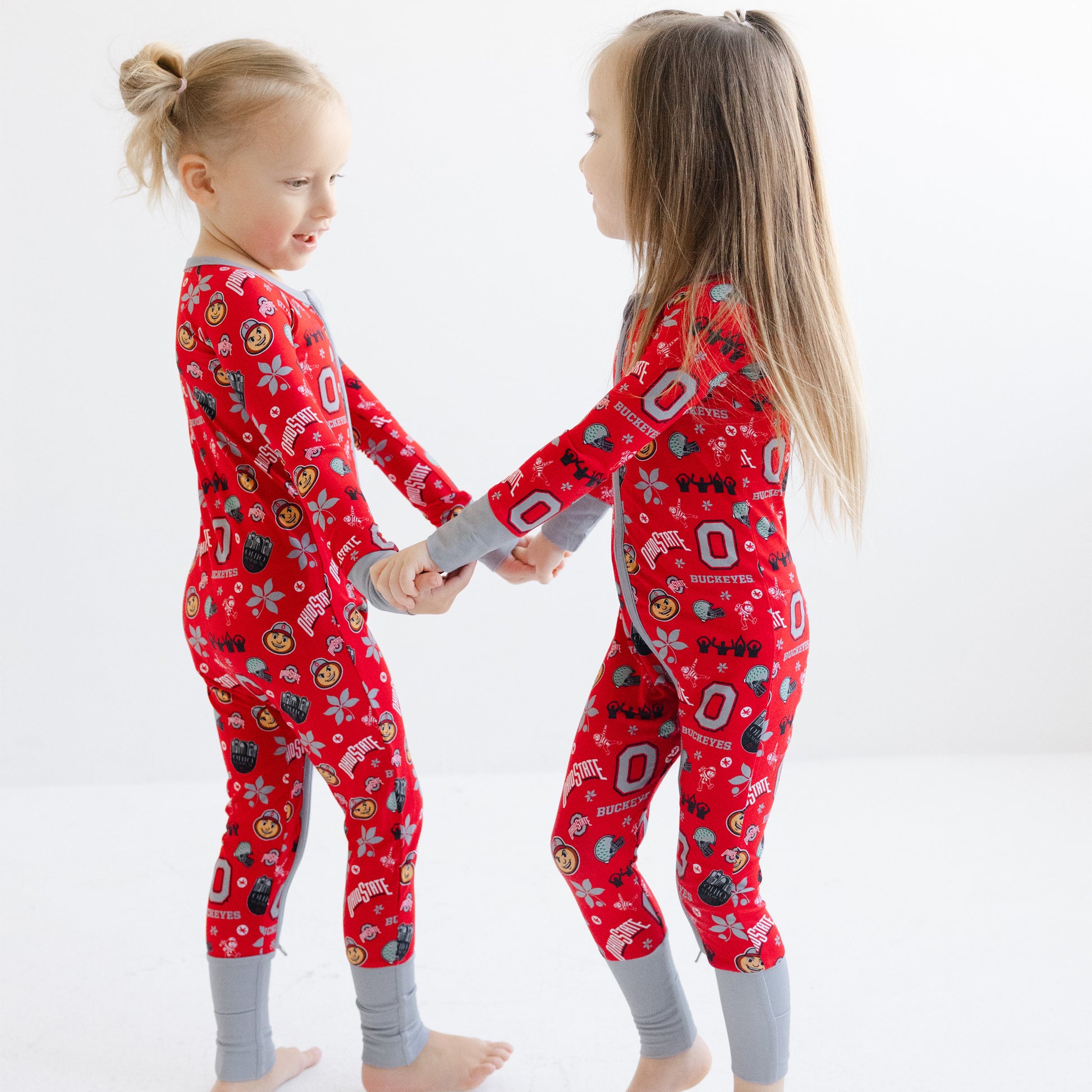 Two young girls standing and holding hands, wearing matching scarlet ohio state buckeyes pajamas with gray cuffs and legs, against a plain white studio background.