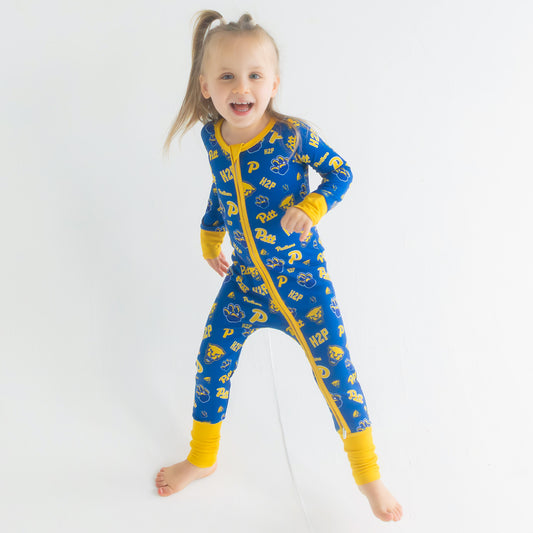 A young girl with blonde hair in a ponytail is smiling and posing in a full-body pajama suit featuring a repeating 'Pitt Panthers' logo pattern, set against a plain white studio background.