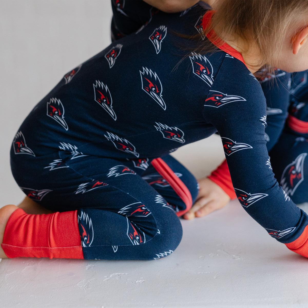 A child wearing a navy UTSA roadrunners bamboo zipper one piece sleeper kneeling on a white surface.