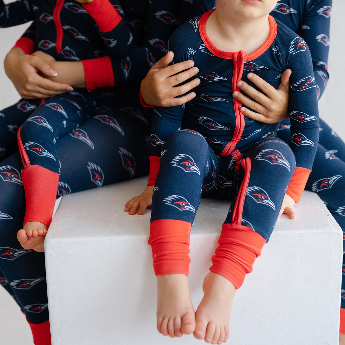 A child wearing a navy UTSA roadrunners bamboo zipper one piece sleeper, sitting on a white block with an adult holding them. The youth wears matching pajamas.