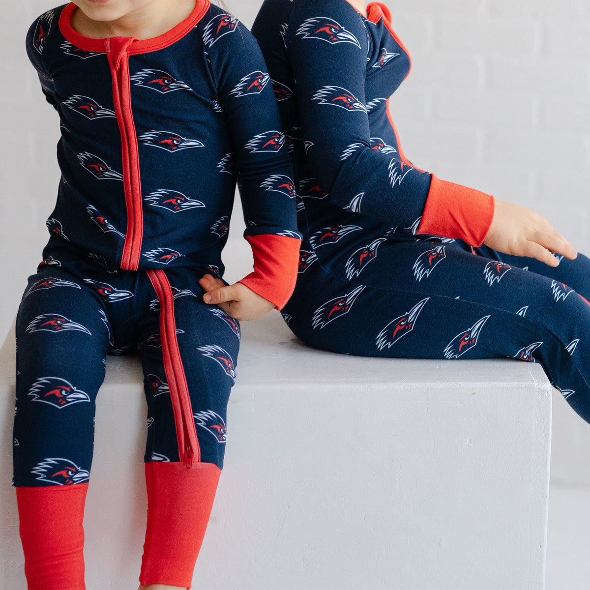 Two children wearing matching navy UTSA roadrunners bamboo zipper one piece sleepers.